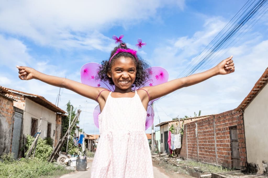 A girl in a pink dress and wings smiles at the camera with her arms raised in victory